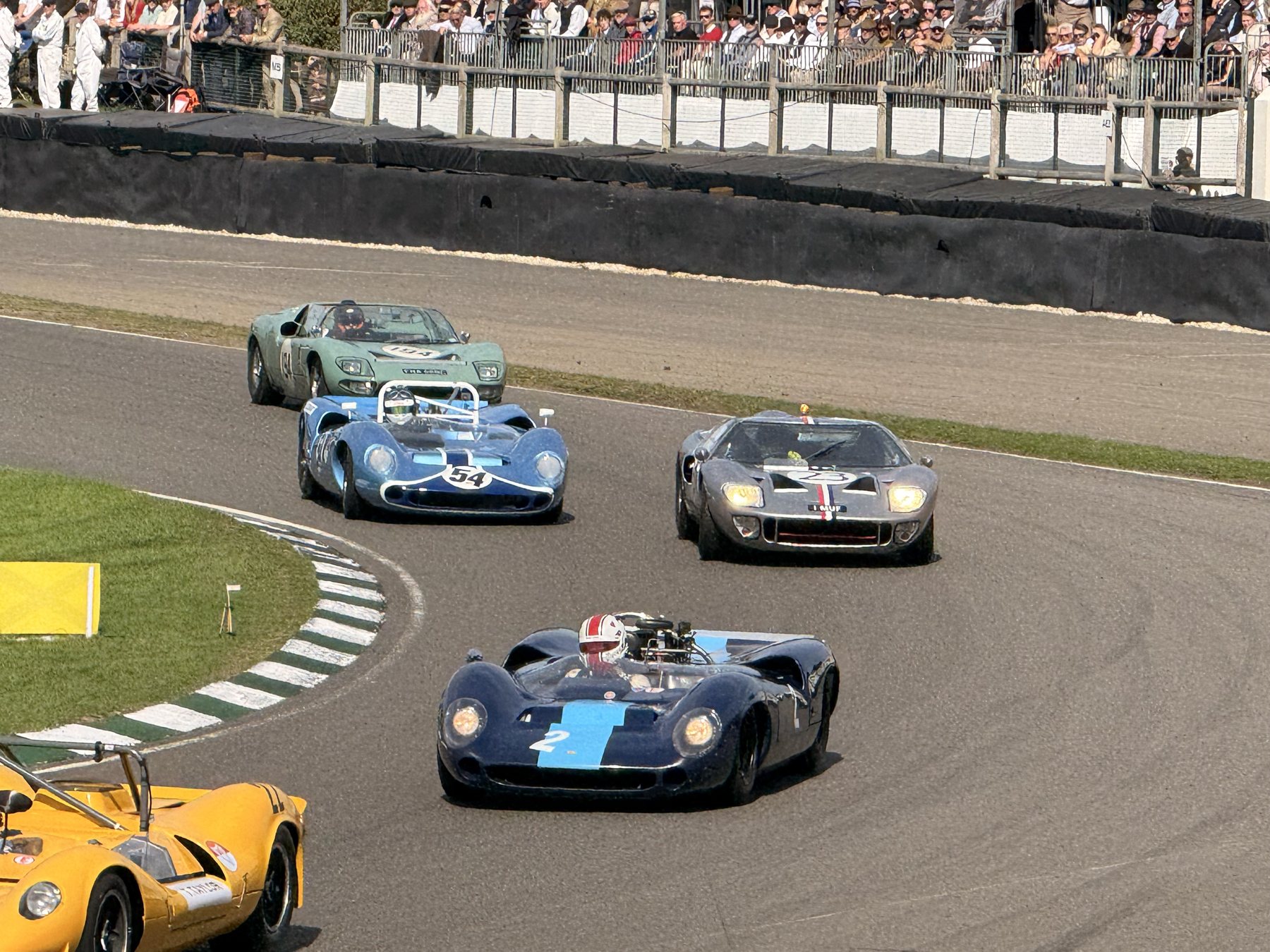 Πακέτο από Lola T70 και Ford GT40 στη σικέιν, με κόσμο στην εξέδρα και ένα κίτρινο αυτοκίνητο στην άκρη