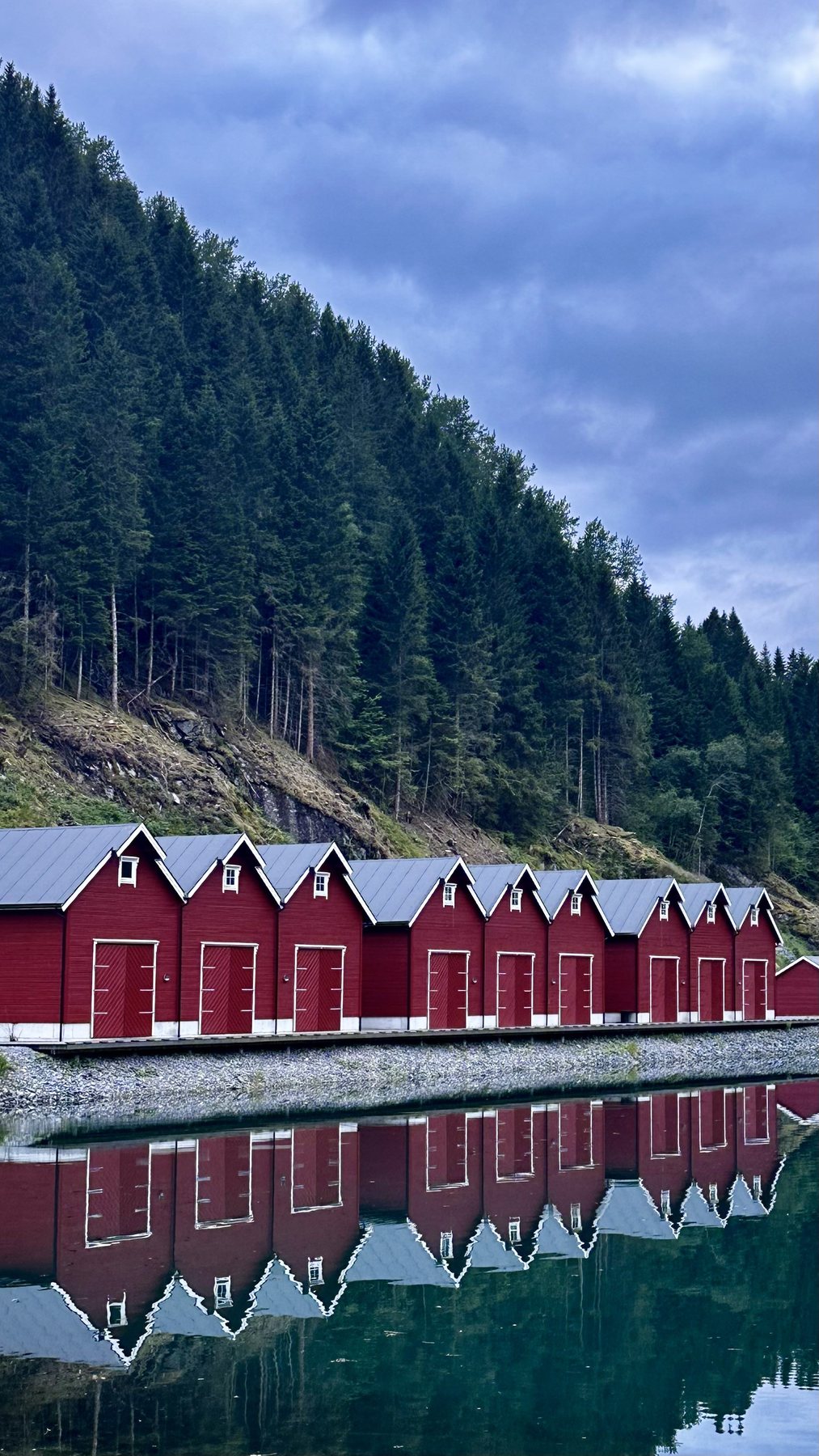 Κόκκινα ξύλινα boathouse καθρεφτίζονται σε ακίνητο fjord, με δάσος από πίσω