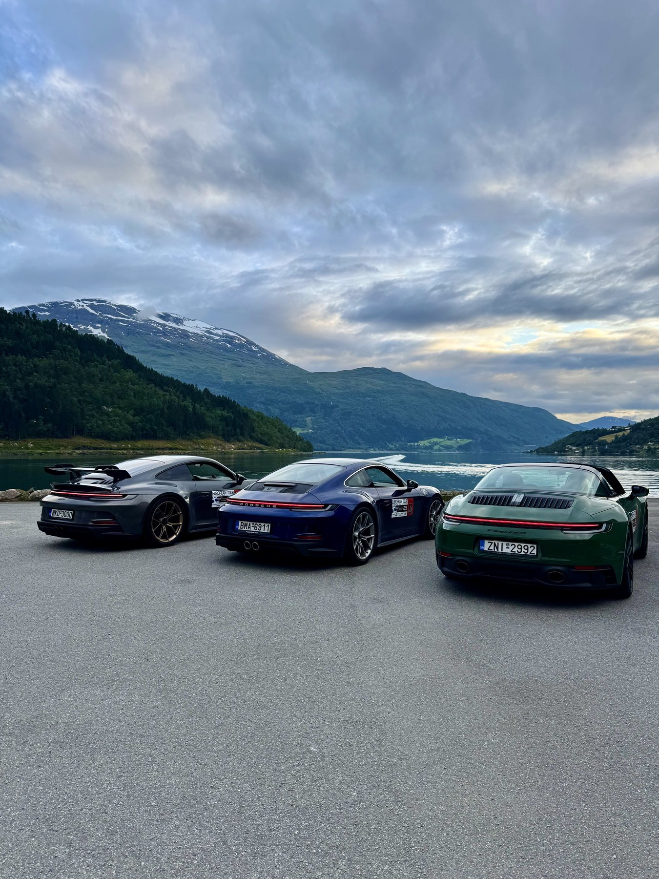 Τέσσερις Porsche παρκαρισμένες σε ένα ξέφωτο δίπλα σε fjord, με βουνά και σύννεφα από πίσω