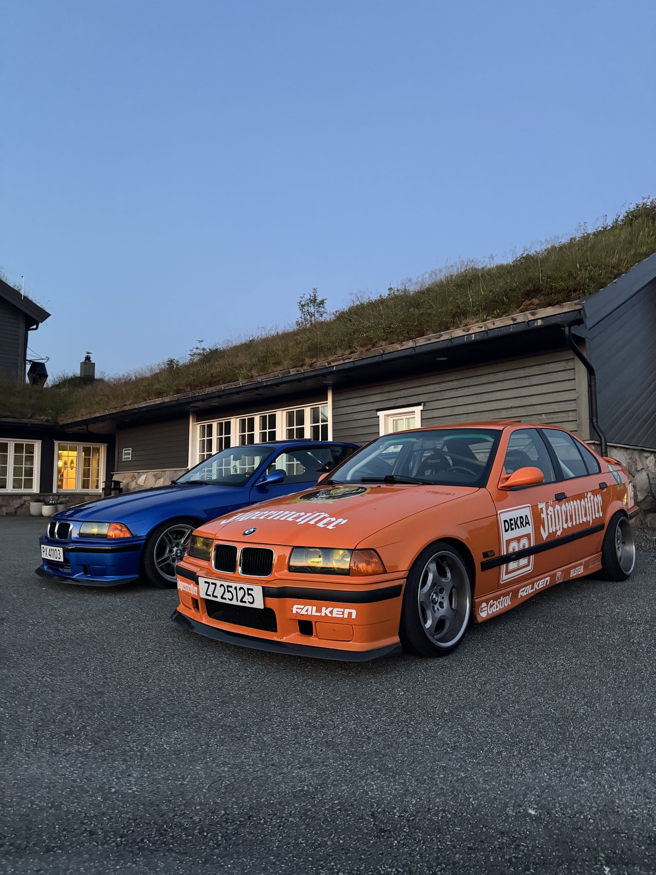 Δύο BMW E36 M3, μία πορτοκαλί και μία μπλε, παρκαρισμένες δίπλα στην 911 μπροστά σε σπίτια με γρασίδι στη στέγη
