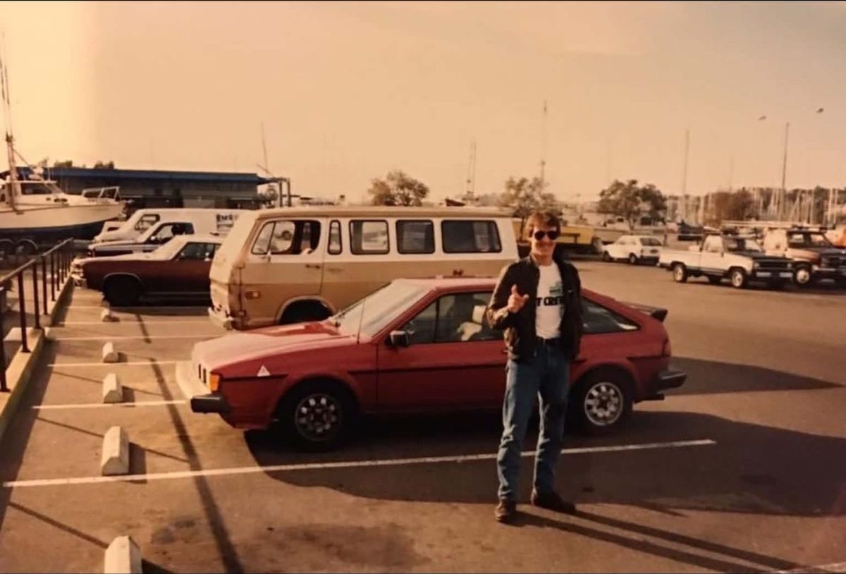 Νεαρός μπροστά από κόκκινο VW Scirocco σε μαρίνα, δεκαετία του ’80