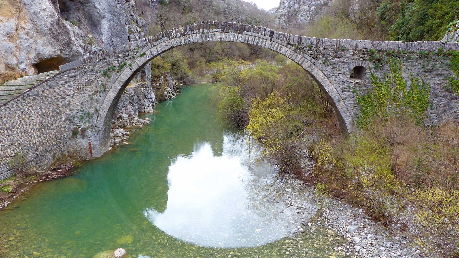 The single-arched Kokkoris stone bridge over green water, between the rocks